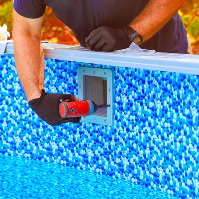 Deux professionnels (homme et femme, 40-50 ans) travaillant ensemble dans l'environnement d'une piscine privée. L'homme en tenue de travail bleu effectue une intervention technique sur un équipement de piscine (filtration ou pompe à chaleur) tandis que la femme, également en tenue professionnelle, l'assiste. Tous deux sont concentrés sur leur travail, montrant une collaboration professionnelle. Arrière-plan : piscine de particulier avec terrasse, végétation, atmosphère sud-française. Image réelle de qualité professionnelle.